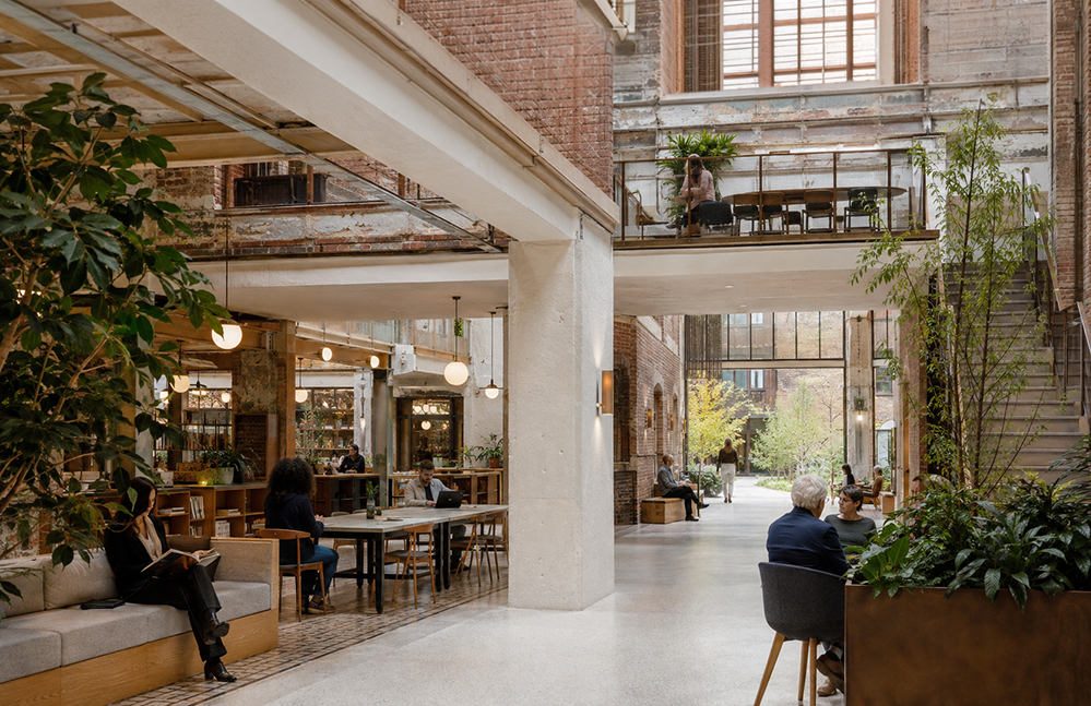 Double-height atrium in a converted brick warehouse used as a co-working space, with people at timber tables, a mezzanine seating area above, globe pendant lights, generous planting and a view through full-height glazing to a planted courtyard. RISE Design Studio.