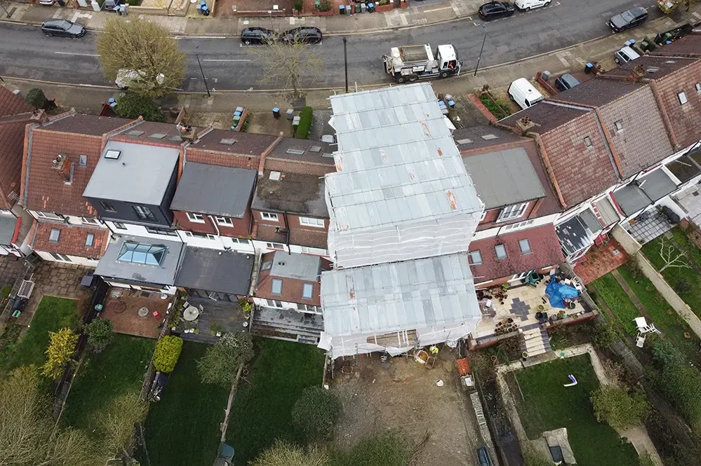 Aerial view of a London terrace mid-renovation, its roof fully wrapped in white sheeting and scaffolding, with a skip and works below, set among neighbouring houses, gardens and rear extensions. RISE Design Studio.