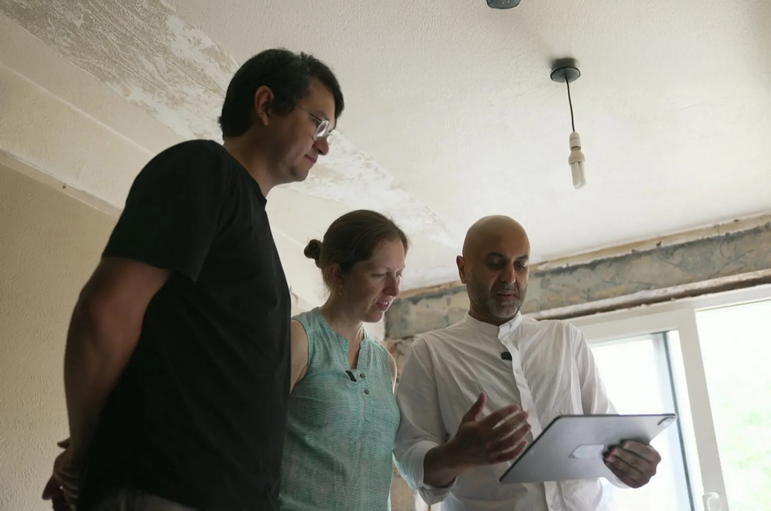 Imran Jahn, sustainability architect stands with two homeowners inside a London house under renovation, reviewing overheating mitigation options using a digital tablet. The space shows stripped-back walls and early retrofit preparation.