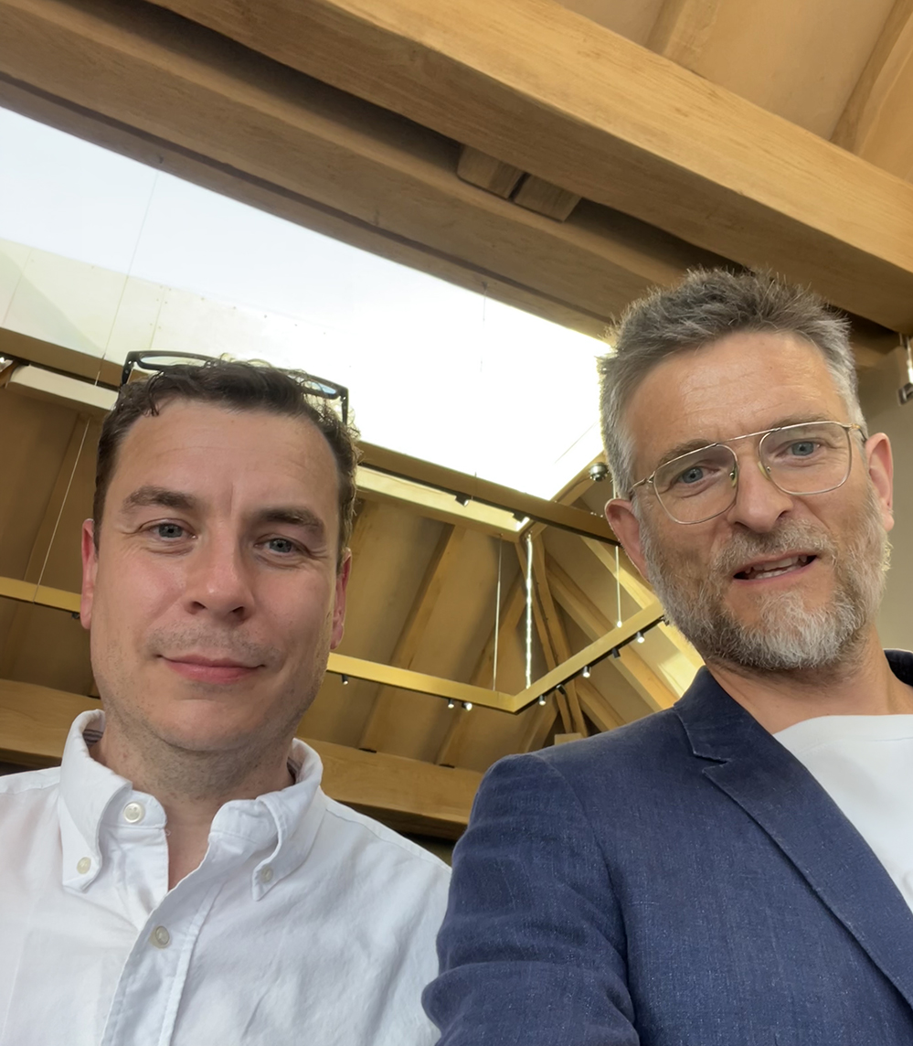 Two men beneath the oak roof structure and glazed rooflight of the Department Store's rooftop level in Brixton, one in a white shirt, one in a navy linen blazer, with the timber frame rising behind them. RISE Design Studio.