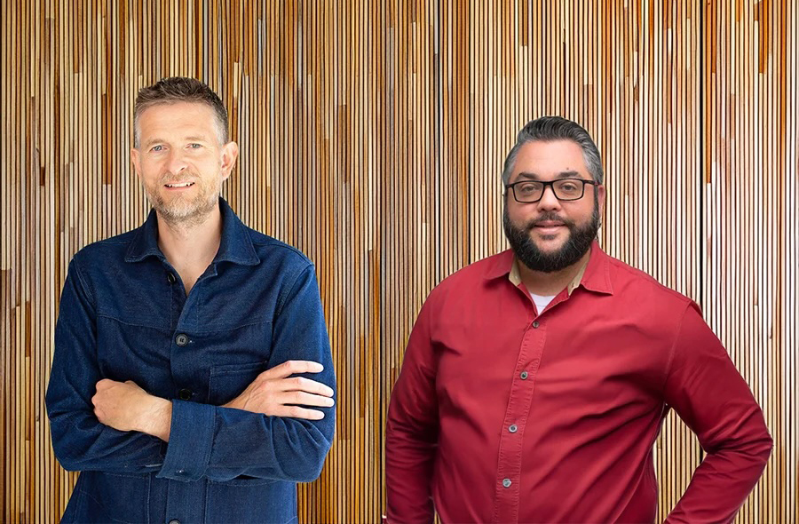 Sean Ronnie Hill (left) and Charlie Cicchetti (right) standing in front of a vertical wooden slat wall. Sean wears a dark blue button-up shirt with arms crossed; Charlie wears a red shirt with glasses and a relaxed smile. Image promoting their podcast discussion on sustainable architecture and green building.