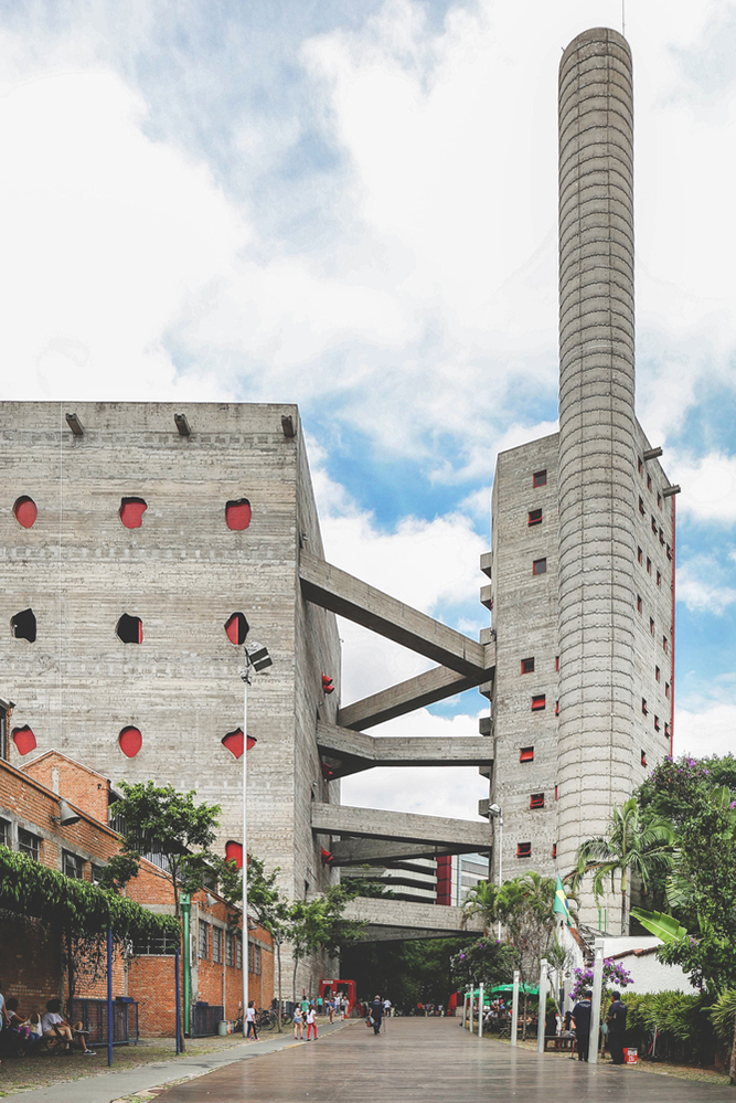 Wide exterior view of Sesc Pompeia’s concrete towers linked by dramatic footbridges, framed by brick buildings and public walkways. The image reveals the strength and clarity of Lina Bo Bardi’s brutalist concrete architecture. Photo by Maria Gonzalez.
